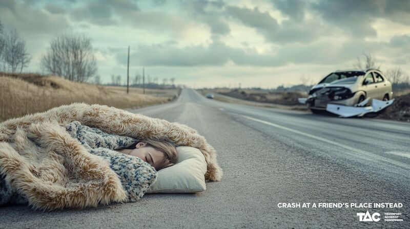 A young woman sleeps on the road in blankets while a crashed car appears in the background.